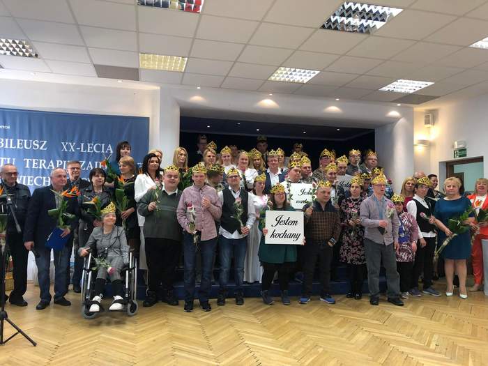 Pokaż zdjęcie: Uczestnicy WTZ w Wiechlicach na tle sceny w trakcie Jubileuszu 20-lecia powstania, z koronami na głowach i kwiatami w rękach