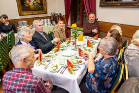 Pokaż zdjęcie: Stół zastawiony na świąteczne przyjęcie, wokół którego siedzi grupa osób w różnym wieku. Na stole ozdobione serwetki, sałatki i dzbanek z sokiem. W tle dekoracje i bufet z kawą oraz ciastem.