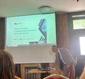 Pokaż zdjęcie: Mężczyzna w garniturze przygotowuje prezentację zatytułowaną „Wsparcie osób z niepełnosprawnością na rynku pracy” wyświetlaną na ekranie. Spotkanie odbywa się w sali konferencyjnej.