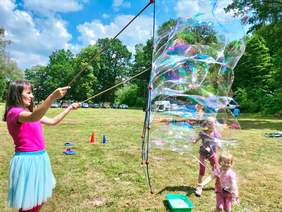 Pokaż zdjęcie: Kobieta puszcza ogromne bańki mydlane, obok stoi dwójka małych uśmiechniętych dzieci