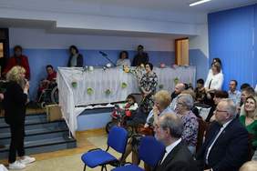 Pokaż zdjęcie: Zdjęcie osób uczestniczących w spotkaniu wielkanocnym, zaproszone osoby siedzą na krzesełkach, kobieta przemawia przez mikrofon. Sala udekorowana jest świątecznie.   