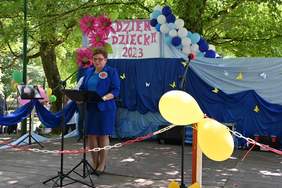 Pokaż zdjęcie: Na scenie w plenerze, stoi kobieta ubrana w niebieski komplet. Scena jest przyozdobiona balonikami.