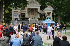 Pokaż zdjęcie: Przed budynkiem kalwarii tańczą w parach uczestnicy festiwalu. Pozostałe osoby siedzą na ławkach.