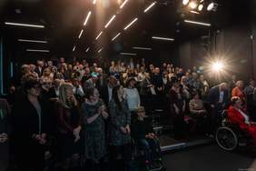 Pokaż zdjęcie: Duża publiczność stoi w sali z ciemnym sufitem i jasnymi liniowymi lampami. Wiele osób bije brawo. Z przodu widoczne są osoby w pierwszych rzędach, w tym dwie kobiety na wózkach oraz osoba w czerwonym stroju po prawej stronie. 