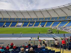 Pokaż zdjęcie: trzy kobiety biegną po niebieskiej bieżni stadionu są dopingowane przez uczestników olimpiady