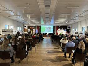 Pokaż zdjęcie: Na dużej sali konferencyjnej na krzesłach w rzędach siedzą ludzie. Z przodu na podwyższeniu jest mównica, za którą stoi mężczyzna. Za nim wielki telebim. Obok banery m.in. baner z logo PFRON.