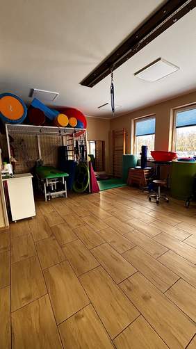 Pokaż zdjęcie: Sala rehabilitacyjna z materacami, piłkami terapeutycznymi, stołem do ćwiczeń oraz drabinkami przy ścianie; pomieszczenie jest jasne i przystosowane do prowadzenia zajęć.