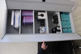Pokaż zdjęcie: Otwarta szafa wypełniona pudełkami w których są klawiatury, słuchawki, aparaty, laptopy