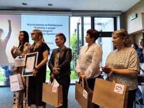 Pokaż zdjęcie: Pięć kobiet stojących koło siebie w rzędzie, pozujących do fotografii. W tle ekran oraz okna sali konferencyjnej.