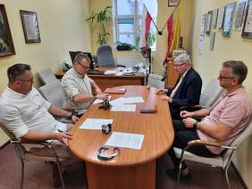 Pokaż zdjęcie: W gabinecie dyrektora znajduje się podłużny stół, przy którym siedzi czterech mężczyzn. Podpisują umowę. Na stole leżą różne dokumenty i pieczątki. W tle widać biurko dyrektora oraz flagi: Polski i Łodzi. 