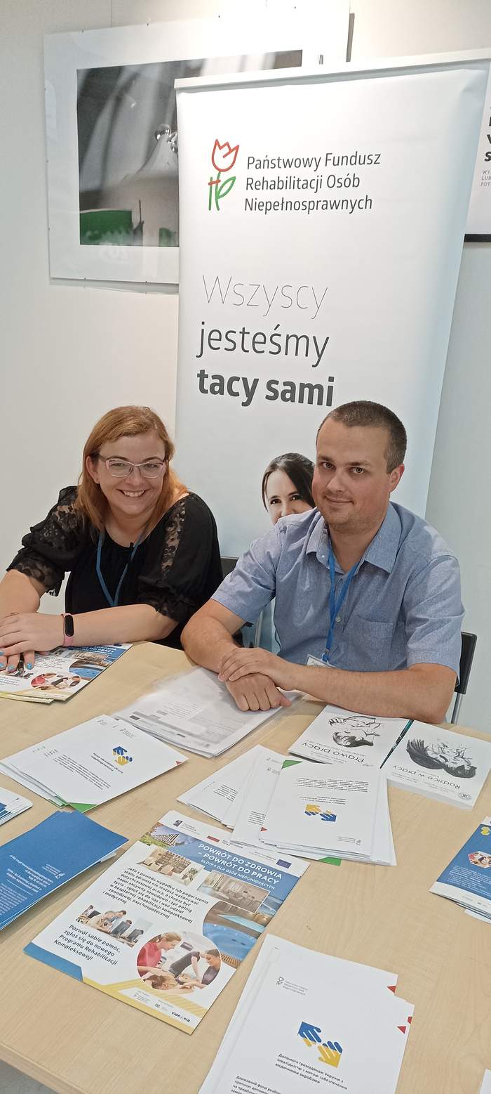 Pokaż zdjęcie: Przedstawiciele Oddziału Lubuskiego PFRON na tle baneru PFRON siedza przy ławce, na której są rozłożone materiały informacyjne