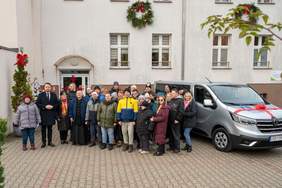 Pokaż zdjęcie: Duża grupa osób, w tym dorosłych i nastolatków, stoi na chodniku przed jasnym budynkiem. Za nimi po prawej stronie zaparkowany jest srebrny van z czerwoną kokardą (taki jak na zdjęciu bus1.jpg). Ludzie są ubrani w ciepłe, zimowe ubrania. Na fasadzie budynku widać dwa wieńce świąteczne.