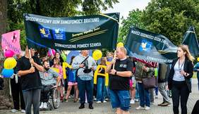 Pokaż zdjęcie: Grupa osób bierze udział w Marszu Godności Osób z Niepełnosprawnościami. Na pierwszym planie widoczne są dwie duże ciemnozielone flagi z nazwami organizatorów: "Środowiskowy Dom Samopomocy Bratanki" oraz "Stowarzyszenie Pomocy Szansa". W tłumie znajdują się osoby na wózkach inwalidzkich, dzieci i dorośli trzymający balony i plakaty.