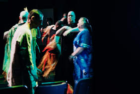 Pokaż zdjęcie: Grupa osób porusza się na scenie teatralnej w dynamicznym układzie; w centrum osoba w ciemnym stroju wykonuje gesty rękami, wokół inni uczestnicy w różnorodnych kostiumach, oświetleni kolorowym światłem, w tle ciemna scena.