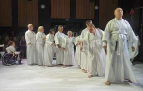 Pokaż zdjęcie: Scena i aktorzy stojący jeden za drugim w białych szatach. Na końcu aktorka poruszająca się na wózku