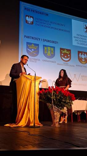 Kobieta przemawia przy mównicy podczas uroczystości jubileuszowej 20-lecia Warsztatu Terapii Zajęciowej w Wałczu. Obok mównicy stoi kosz z czerwonymi różami. W tle wyświetlana jest prezentacja dotycząca jubileuszu, a na scenie widoczne są materiały promocyjne warsztatu.