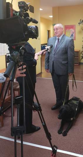 Pokaż zdjęcie: pomieszczenie Oddziału korytarz po prawej stronie stoi osoba w ręku trzyma smycz  z psem przewodnikiem, po lewej stronie kamera 