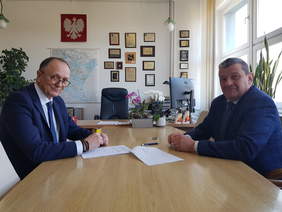 Dwóch mężczyzn w garniturach siedzi przy stole konferencyjny. Odwróceni w kierunku robiącego zdjęcie.