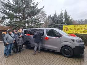 Pokaż zdjęcie: Przy samochodzie stoi grupka osób. Pojazd jest udekorowany czerwoną wstążką. W tle widać plakat Domu Pomocy Społecznej Fundacja im. Brata Alberta w Łodzi. 