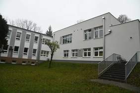 Pokaż zdjęcie: Zdjęcie jednopiętrowego budynku w jasnym kolorze, w kształcie litery L.