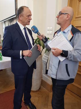 dwóch mężczyzn, stoją, jeden udziela wywiadu drugiemu, do mikrofonu