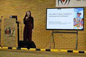 Pokaż zdjęcie: Kobieta stoi na scenie z mikrofonem w ręku i prowadzi prezentacje na temat „Jak radzić sobie z trudnymi zachowaniami małego dziecka?”.  W tle widoczny ekran z wyświetlana prezentacją.