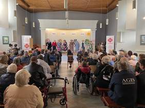 Pokaż zdjęcie: W oddali scena na której stroją uśmiechnięte seniorki w kapeluszach i wzorzystych kolorowych sukienkach. Trzymają się za ręce. Na sali siedzi dużo gości oglądających spektakl. Są też osoby na wózkach inwalidzkich.