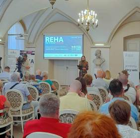 Pokaż zdjęcie: Grupa około dwudziestu osób siedzi w sali konferencyjnej na eleganckich krzesłach. Z przodu znajdują się: kamerzysta, osoba przemawiająca do mikrofonu, duży ekran z wyświetloną prezentacją, jak również banery organizatorów wydarzenia oraz PFRON.