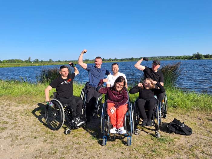 Pokaż zdjęcie: Nad jeziorem, na tle bezchmurnego nieba znajduje się grupa kilku osób poruszających się na wózku. Większość osób, w geście zwycięstwa, ma uniesione w górę ręce.