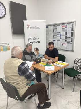 Pokaż zdjęcie: Starszy mężczyzna siedzi przy stoliku i rozmawia z pracownikami podczas konsultacji w ramach dyżuru eksperckiego PFRON. Na stole leżą dokumenty i materiały informacyjne, a w tle widoczny jest roll-up PFRON z hasłem „Wszyscy jesteśmy tacy sami”.