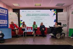 Pokaż zdjęcie: Pięć kobiet bierze udział w panelu dyskusyjnym na konferencji ZUS. Cztery panelistki siedzą na czerwonych fotelach, a jedna uczestniczka porusza się na wózku elektrycznym i mówi do mikrofonu. W tle duży ekran z tytułem panelu i nazwiskami prelegentek, banery ZUS i PFRON. Na stoliku stoją butelki wody. W lewym dolnym rogu widoczny jest ekran z tłumaczem języka migowego.