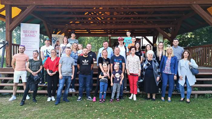 Pokaż zdjęcie: Uczestnicy pikniku z okazji Międzynarodowego Dnia Głuchych. Grupa uczestników stoi na tle drewnianej sceny