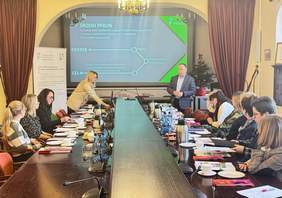 Pokaż zdjęcie: Sala konferencyjna z długim drewnianym stołem i ludźmi siedzącymi po obu jego stronach. Na środku stołu leżą liczne dokumenty, notesy i kolorowe broszury, stoją butelki wody, termosy z napojami, szklanki i małe talerzyki. Przy miejscach uczestników spotkania znajdują się mikrofony konferencyjne. Po obu stronach stołu siedzą osoby w eleganckich strojach, pochylone nad materiałami lub zwrócone w stronę przodu sali.   Przy przednim końcu stołu stoi kobieta w jasnym żakiecie, pochylona nad laptopem i prowadzi prezentację. Obok niej, z prawej strony przy ekranie projekcyjnym, stoi mężczyzna w ciemnej marynarce i jasnej koszuli. Na ścianie wyświetlona jest prezentacja w odcieniach zieleni i błękitu z dużym napisem „Środki PFRON” oraz słowami „oferta” i „cel”, a po prawej stronie widnieje logo Państwowego Funduszu Rehabilitacji Osób Niepełnosprawnych.