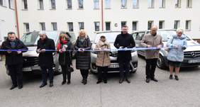 Pokaż zdjęcie: cztery kobiety i czterech mężczyzn stoją w rzędzie w dłoniach trzymają nożyczki, przecinają wstęgę. za nimi stoją trzy duże busy.