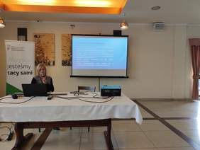 Pokaż zdjęcie: Przy stole ma którym jest laptop siedzi kobieta w w dłoniach trzyma mikrofon. Za nią na ekranie wyświetlona jest prezentacja.