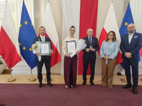 Pokaż zdjęcie: Pięć osób laureatów  konkursu pozuje z dyplomami na tle flag Polski i Unii Europejskiej.