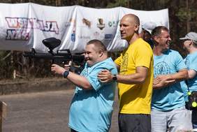 Pokaż zdjęcie: Uczestnik Spartakiady trzyma karabin paintballowy, celuje do celu pod okiem instruktora. Za nimi stoją uczestnicy zawodów sportowych. W tle widać banery sponsorów i innych uczestników.