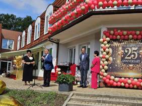 Pokaż zdjęcie: Mężczyzna w garniturze i kobieta stoi przed udekorowanym budynkiem z okazji 25-lecia. Balony w złoto-różowych kolorach, baner z napisem “Happy 25th Anniversary”