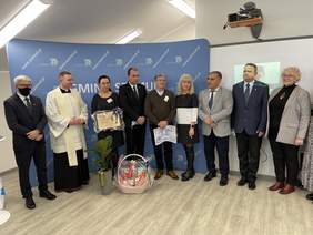Pokaż zdjęcie: Kilka osób ubranych na galowo wraz z księdzem stoi w jednym rządzie. Przed nimi na podłodze leży kosz upominkowy, jedna z kobiet trzyma w ręku tablicę z grawerem. Za nimi stoi szeroki niebieski baner z napisami Gmina Szemud.