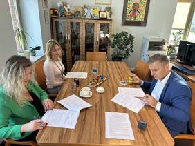 Pokaż zdjęcie: Przy drewnianym dużym stole siedzą trzy osoby, dwie kobiety w blond włosach po lewej stronie i mężczyzna w niebieskiej marynarce, białej koszuli po prawej stronie. Podpisują dokumenty. Za nimi komoda na której stoją kolorowe pamiątki. Po prawej 