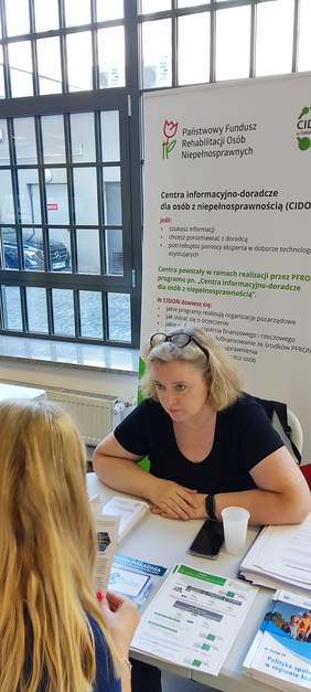 Pokaż zdjęcie: sala NFZ pracownik odddziału siedzi przy stole na stoisku i udziela porad i informacji na temat dofinansowań ze środków PFRON