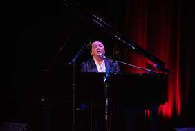 Pokaż zdjęcie: Na scenie przy fortepianie siedzi mężczyzna w ciemnym garniturze i śpiewa do mikrofonu