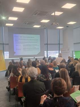 Pokaż zdjęcie: kobieta siedząca za stołem prezydialnym w Sali konferencyjnej, na ekranie wyświetla się prezentacja