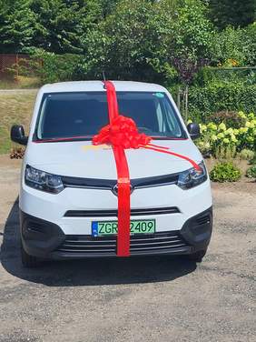 Pokaż zdjęcie: Przód białego, dostawczego auta z czerwoną kokardą na masce.