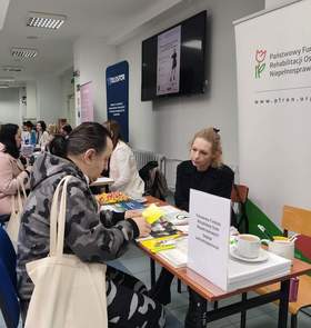 Zdjęcie przedstawia stoisko PFRON w trakcie trwania wydarzenia. Przy stoliku siedzi kobieta (przedstawicielka instytucji) w czarnym ubraniu, która rozmawia z mężczyzną siedzącym naprzeciwko niej. Mężczyzna w bluzie moro przegląda żółtą ulotkę. Na stoliku, obok materiałów promocyjnych i słodyczy, stoi biała filiżanka z kawą. W tle widać długi korytarz z innymi stoiskami targowymi (m.in. firmy Telesfor), przy których siedzą ludzie. Na ścianie powyżej stoiska zawieszony jest ekran telewizyjny wyświetlający plakat informacyjny.