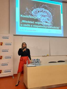 Pokaż zdjęcie: W auli sali wykładowej stoi kobieta, która przemawia do mikrofonu. Nad nią na tablicy multimedialnej wyświetlony jest temat konferencji. 