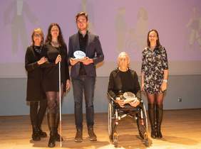 Pokaż zdjęcie: na zdjęciu scena na której stoi 5 osób w tym jedna siedzi na wózku inwalidzkim, druga trzyma w ręku białą laskę oraz trzymają statuetki z napisem pełnia życia 