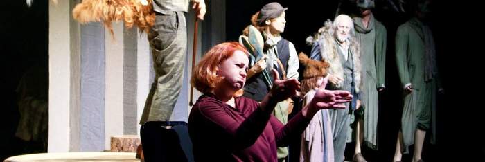 Pokaż zdjęcie: Przed sceną stoi kobieta w czarnej bluzce z rękami wyciągniętymi do przodu. Za nią na scenie stoją aktorzy.