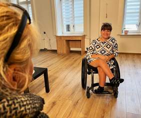 Pokaż zdjęcie: Na środku pomieszczenia na wózku inwalidzkim siedzi młoda dziewczyna. Jest ubrana w śliczną sukienkę i uśmiecha się szeroko. 