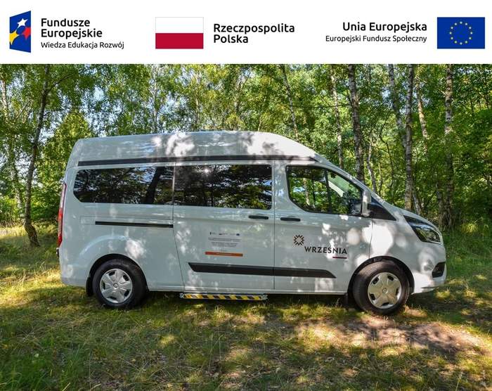 Pokaż zdjęcie: Na górze, od lewej strony widoczne są następujące logotypy: Funduszy Europejskich – Programu Wiedza-Edukacja-Rozwój; Rzeczpospolitej Polskiej oraz Unii Europejskiej – Europejskiego Funduszu Społecznego. Poniżej na polanie, w pobliżu zagajnika brzozowego stoi biały bus z logotypem Gminy Września oraz naklejką informującą o dofinansowaniu zakupu pojazdu w ramach konkursu grantowego PFRON pn. „Usługi indywidualnego transportu door-to-door oraz poprawa dostępności architektonicznej wielorodzinnych budynków mieszkalnych”.